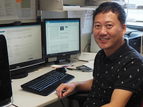 A man in a black polo sits at a desk in front of two computers.