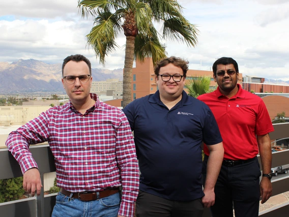 Three men pose for a photo on a rooftop. A palm tree and the Tucson mountains lay in the background.