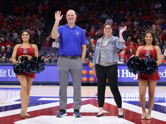 four people stand and wave from a basketball court.