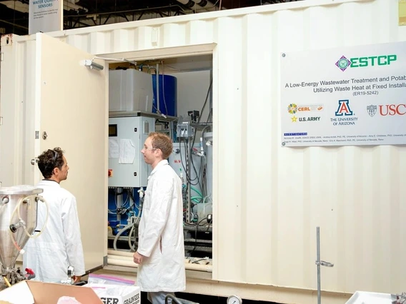 Two men in lab coats stand in front of a large metal shipping container.
