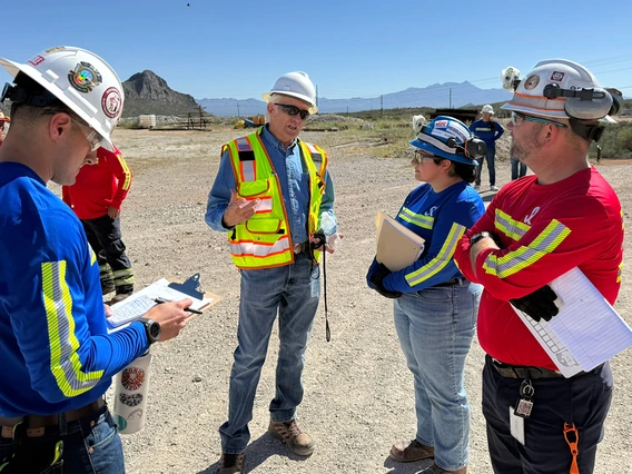 Five people in yellow vests stand on dusty land.