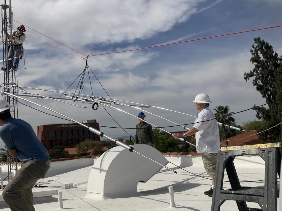 Five people on a roof installing an antenna and wearing hard hats. Four are on the surface of the roof while one is climbing up a metal structure.