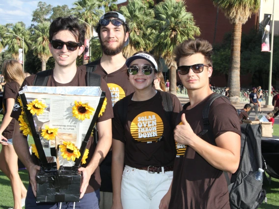 Four students gather around their solar oven, which is made of cardboard and foil and decorated with plastic sunflowers