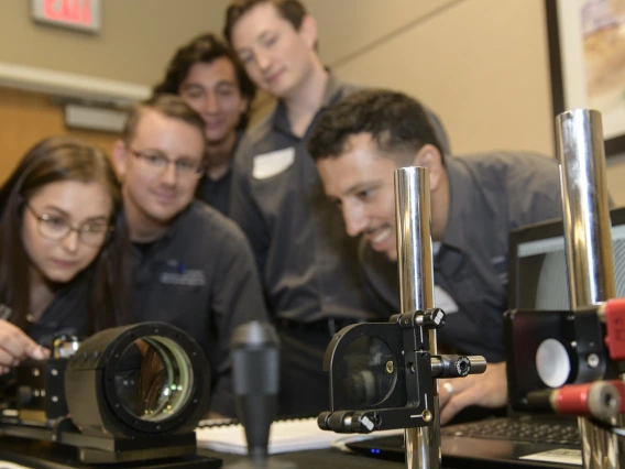 Five students lean over a display of optical equipment