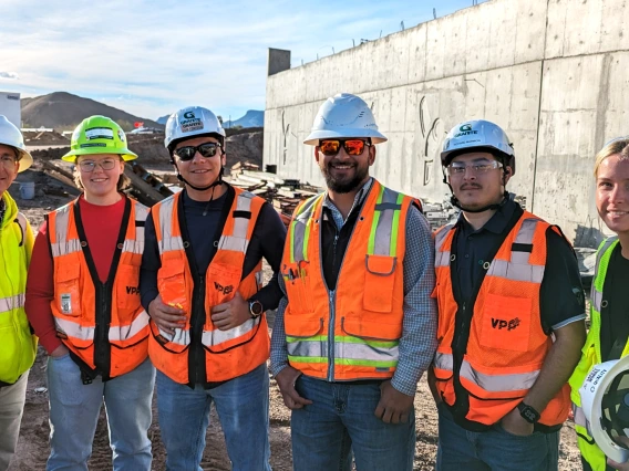 Six people pose outdoors at a construction site in hard hats