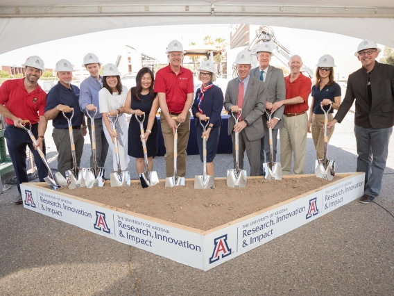 A group of a dozen people wearing white construction hats hold shovels that are placed into a square of dirt.