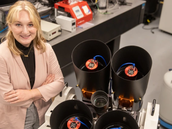 A young woman wearing a blazer is crossing her arms and smiling. She's standing in a lab environment, and next to her are four black cylinders, approximately 10 inches in diameter, win imaging instrumentation inside.