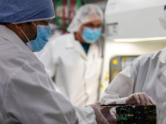 Two masked students work in a cleanroom with satellite components