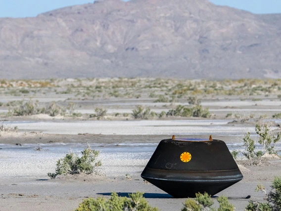 A black metal capsule the size of a doghouse rests on the desert ground