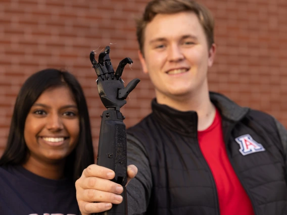 Nisha Rajakrishna and Collin Preszler in the background. Collin is holding up a black prosthetic arm in the foreground.