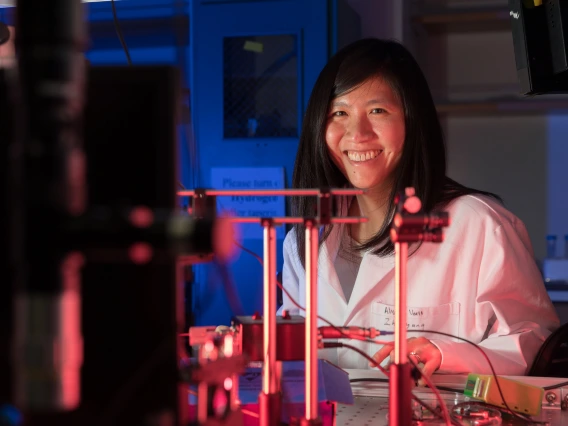 Judith Su wearing a white labcoat and smiling behind a complicated array of sensors.