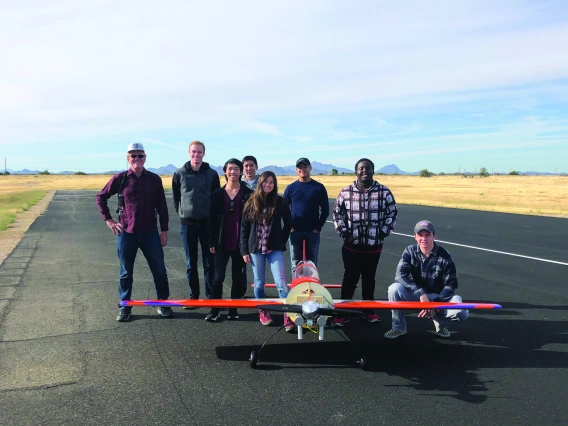 A group of eight people stand smiling for a photo with a hand-built airplane, which sits in front of them and looks to have a wingspan of about 10 feet.