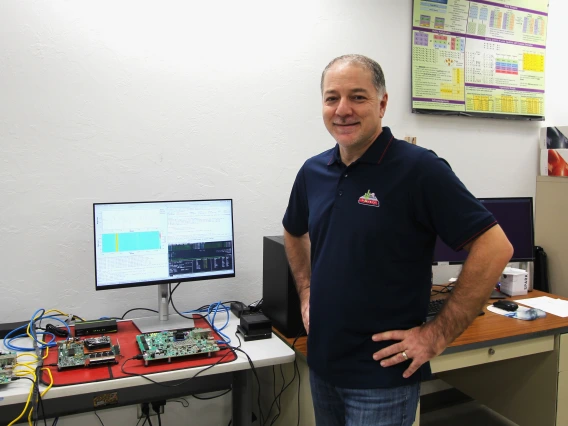 Ali Akoglu in his lab. His hands are on his hips and he is smiling. Behind him is a computer monitor and a green circuit board.