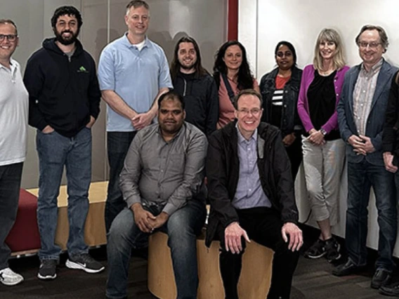 Team members pose in a room with a glass wall and whiteboard