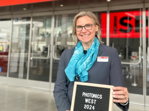 A woman in a blue scarf, Jennifer Barton, holds a sign that reads Photonics West 2024.