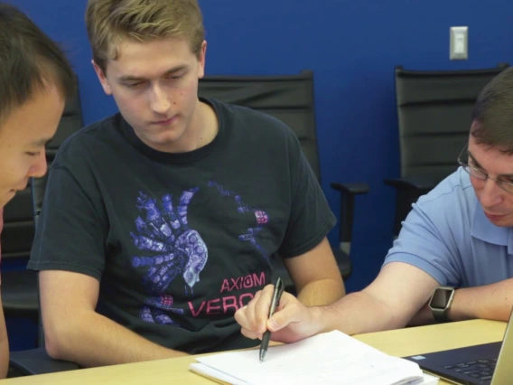 Three men lean over a sheet of paper and a laptop, the one on the far right pointing and explaining a concept.