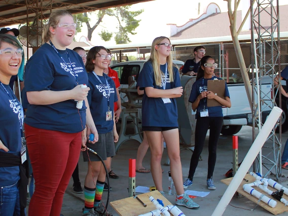 A group of high school girls wearing matching blue "Summer Engineering Academy" t-shirts grin off into the distance at a potato (off frame) they just launched out of a homemade potato launcher.