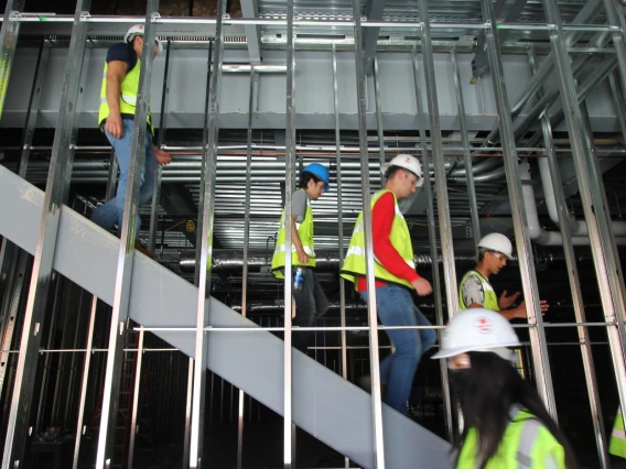 A group of students in yellow vests and hard hats walk down a set of stairs behind a set of beams