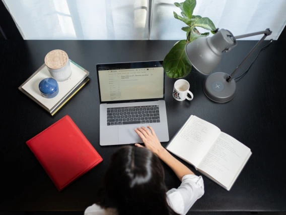 A photo of a woman at a desk, taken from overhead. the desk has a laptop, an open notebook, a stack of folders, a plant, a lamp and a University of Arizona coffee mug.