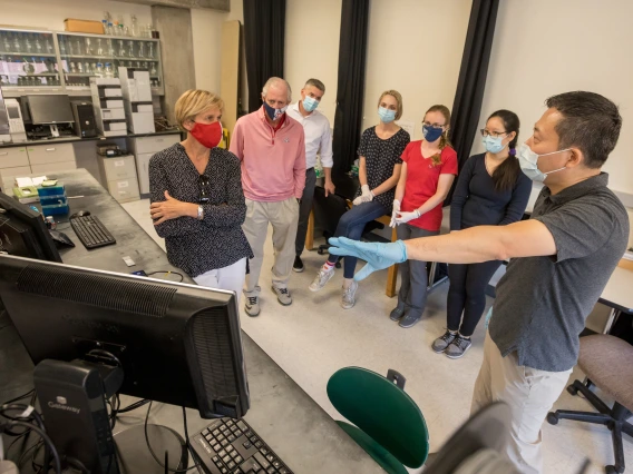 A man gestures to a computer on a laboratory counter, showing a small group of people something on the screen.