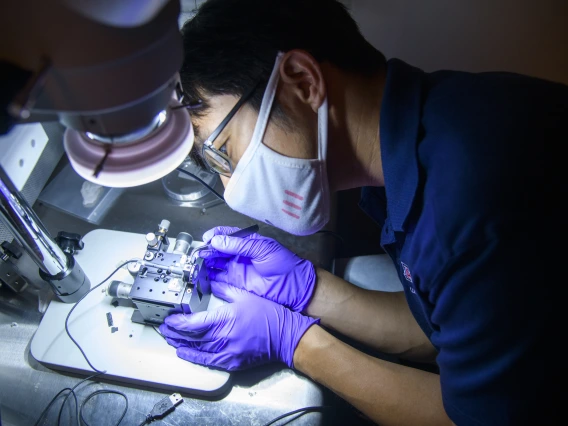 Dongkyun Kang, wearing rubber gloves and a mask, leans over to work on a small device under a bright light.