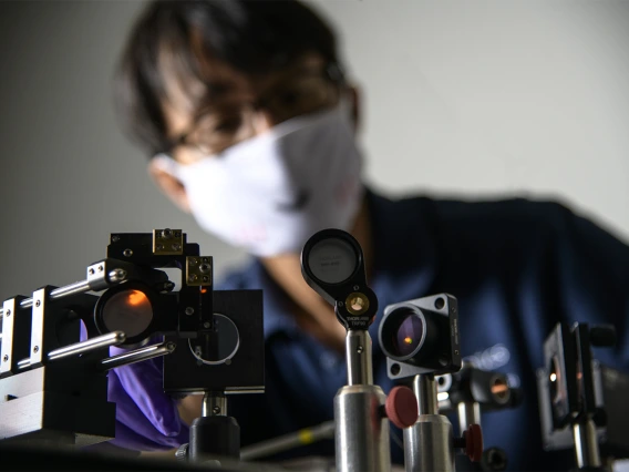 A set of optical devices in the foreground, with DK Kang in the background, blurred and wearing a white face mask.