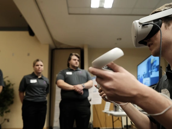 A student seen from the side wearing a VR headset and holding VR hand controllers, while students in the background look on.