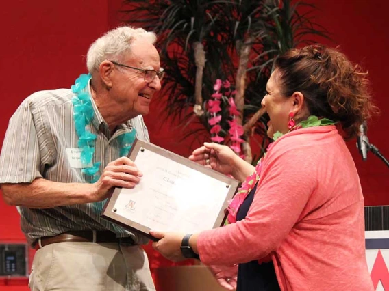 William Cosart, wearing a blue lei, hands a framed certificate to Claudia Arias.