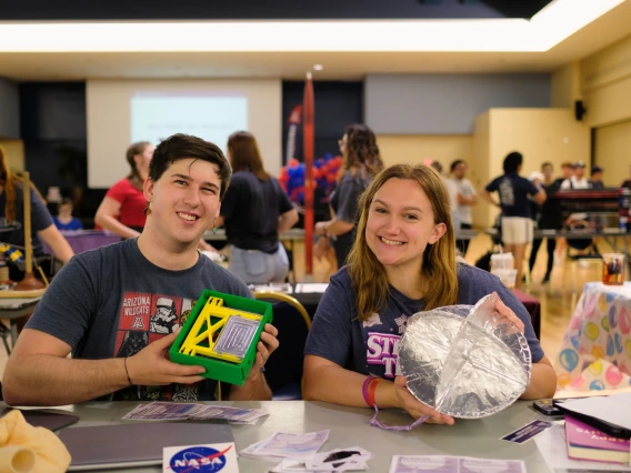 Two students sit at a booth for the Engineering Club showcase and smile. He is holding a green and yellow box and she is holding a circular metallic object..