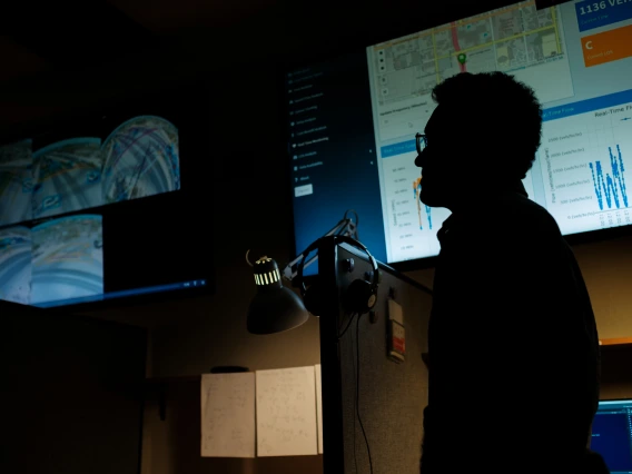 A silhouette of a man wearing glasses in front of a large computer screen.