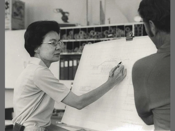 Frances Walker explaining a blueprint on a large white sheet of paper to an onlooker.