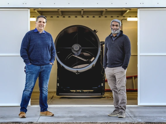 Roberto Furfaro and Vishnu Reddy stand in front of a piece of telescope equipment, a cylinder about 3-4 feet in diameter.