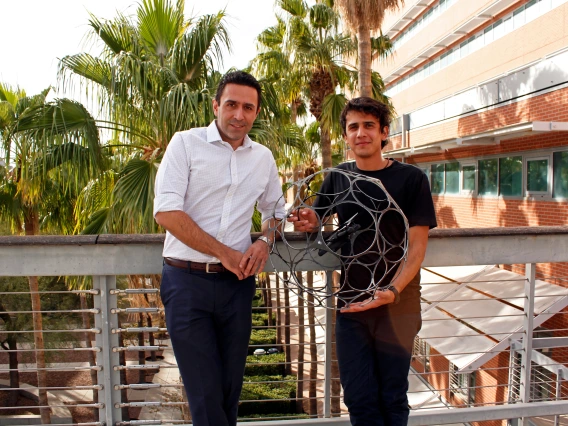 Two men stand in front of a railing and some palm trees, holding a drone encased in a spherical cage, about three feet in diameter