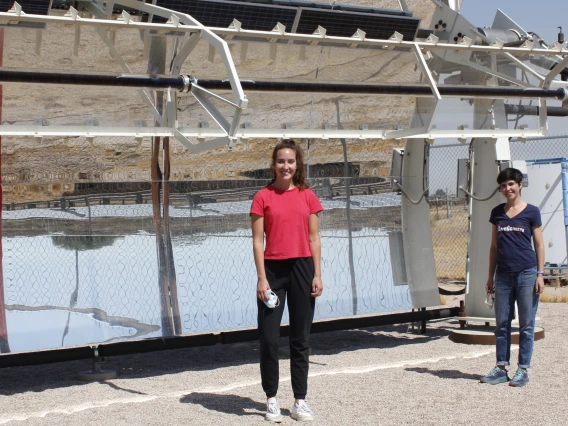 Two women stand in front of a solar-powered desalination system, shaped like a giant curved mirror