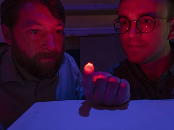 Two men, visible from the shoulders up, in a dimly lit room. The man on the left is holding a small, reddish light source on his hand, which is illuminating their faces. Both are staring at the light.