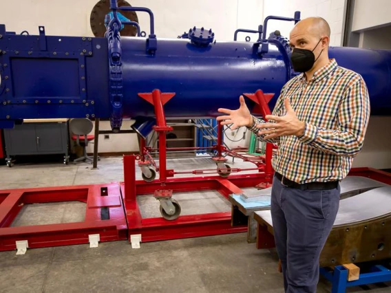 Jesse Little stands in front of a large blue wind tunnel -- it looks like a horizontal pipe -- on red scaffolding. He is wearing a black mask over his nose and mouth and gesturing with his hands to explain something.