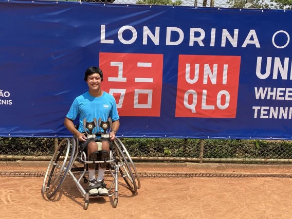 Jason Keatseangsilp sits in a sports wheelchair with slanted wheels, smiling and holding two trophies on his lap. Behind him is a blue banner that reads, "Londrina Open UNIQLO Wheelchair Tennis Tour"