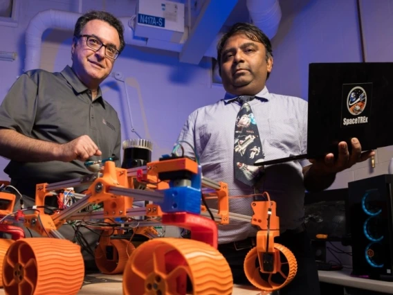 In the foreground, a robot with four orange wheels. Two men stand behind the robot.