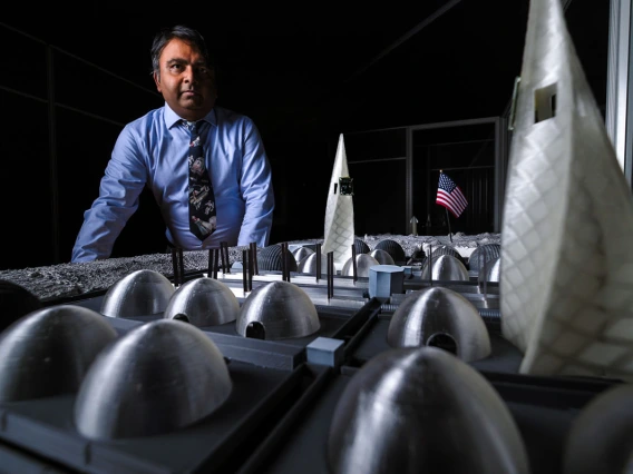 A man in a blue shirt stands in front of a small-scale table that mimics a lunar base.