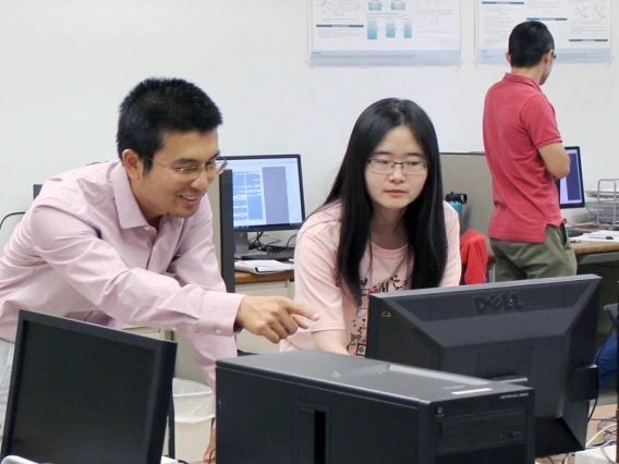 Ming Li leans over a computer, smiling and helping a student.