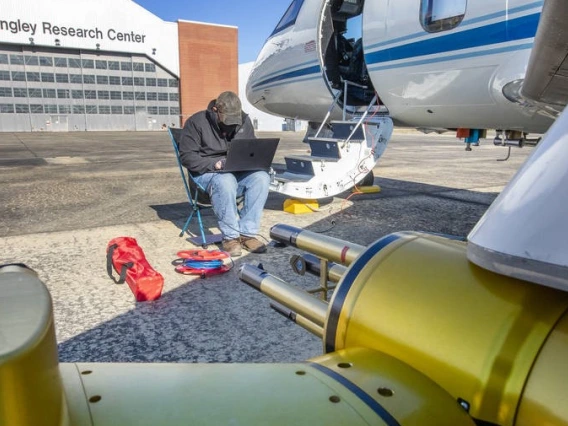 A man sits on a folding chair using a laptop, next to the steps of an airplane. The building behind him has a sign that says, "Langley Research Center."