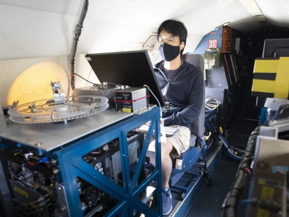 A young man wearing a face mask sits behind a monitor, surrounded by technology on a plane.