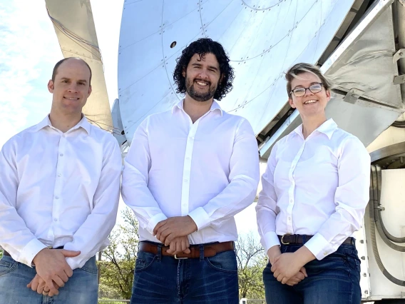 Three people in white shirts stand in front of a large, shiny telescope.