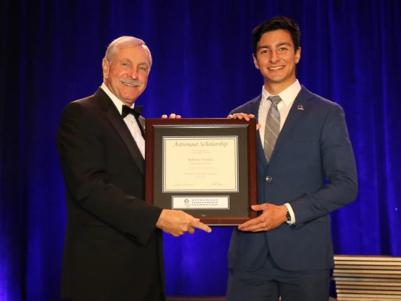 Roberto Peralta stands on a stage accepting a framed certificate from an older man. The certificate says "Astronaut Scholarship." The men are in formal wear and there is a blue curtain behind them.