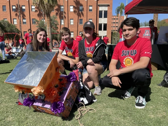 Four students in matching red t-shirts pose on the lawn next to their solar oven -- a cardboard box with a funnel shape on top, decorated with orange and purple tissue paper.