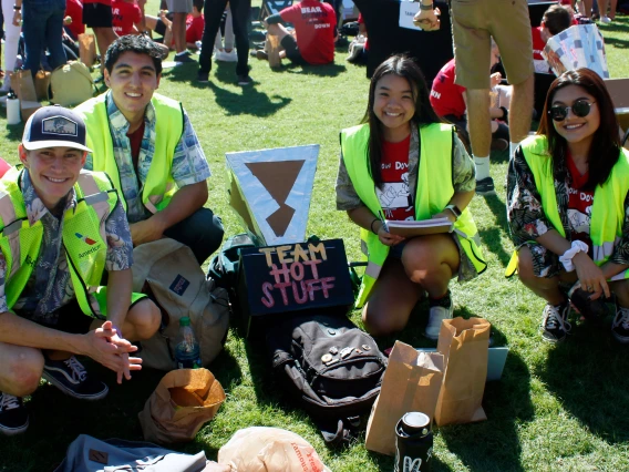 Two young men and two young women wearing Hawaiian shirts and bright yellow vests kneel for a photo next to their solar oven, a cardboard and aluminum foil device labeled with their group name: "Team Hot Stuff."