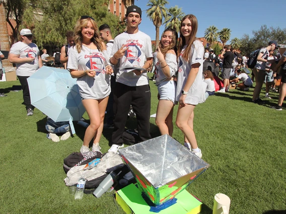 Four students in white shirts smile and pose for a photo on a grassy lawn with an oven made of aluminum foil and cardboard at their feet.