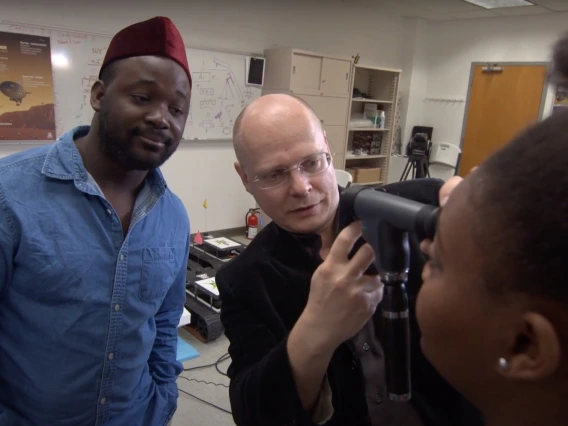 Wolfgang Fink uses an optical device to examine a young woman's eyes, while another young man looks on in the background.