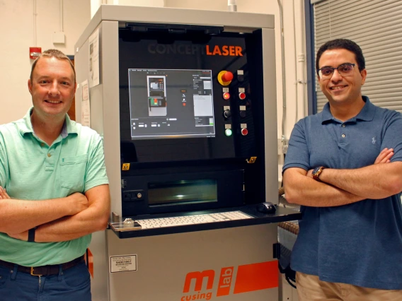 Andrew Wessman and Mohammed Shafae standing next to a 3D printer, which is about two feet wide and a bit taller than them.