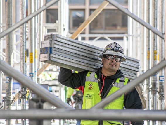 A man wearing a construction vest carries four metal beams on a construction site.